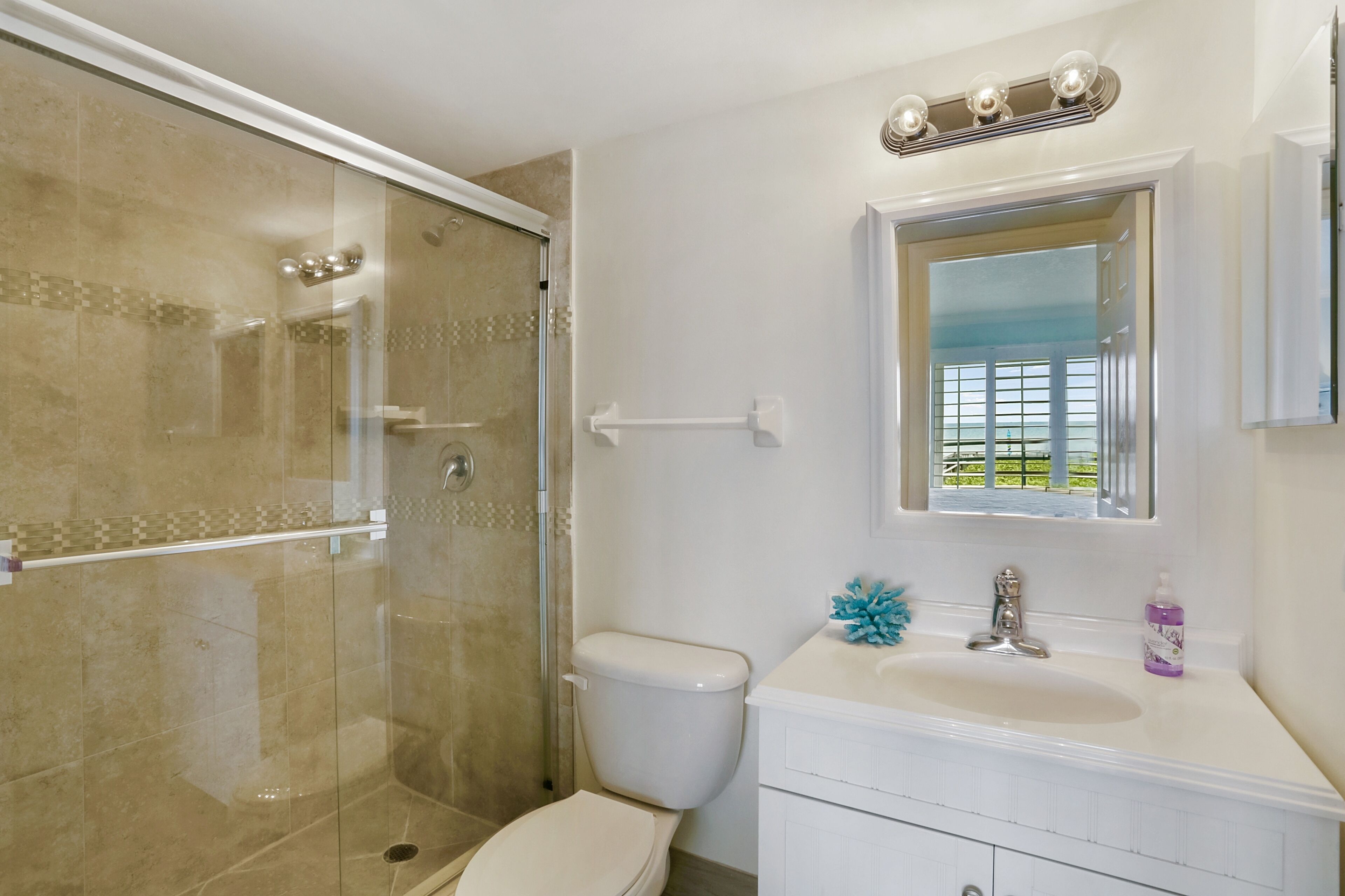 A bathroom with a glass-enclosed shower, a toilet, a sink with a mirror above it, and a window with a view of greenery.