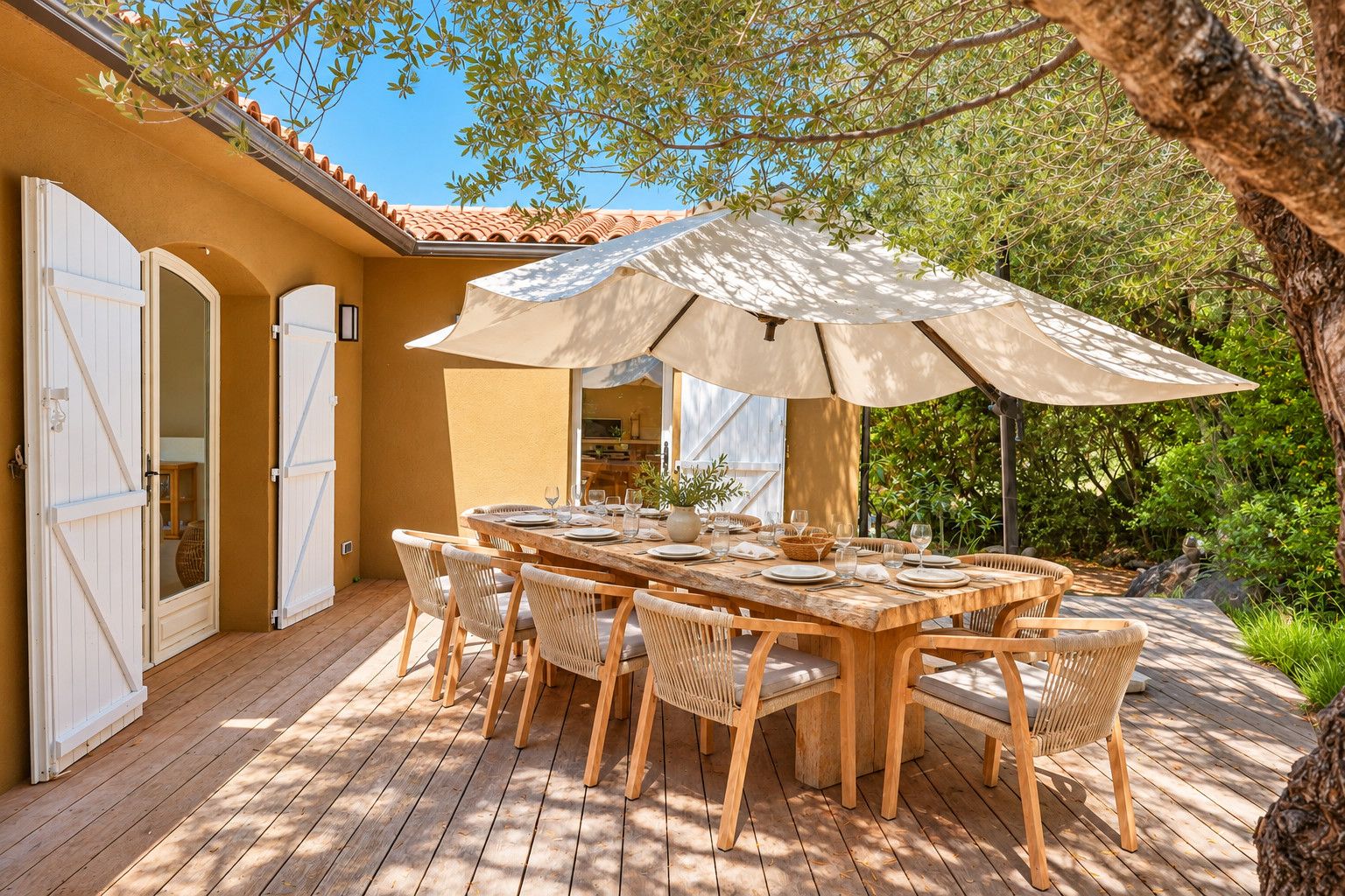 Un área de comedor al aire libre con una mesa y sillas de madera, un paraguas grande y vistas a un jardín con árboles y un edificio al fondo.
