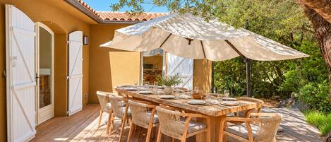 Un área de comedor al aire libre con una mesa y sillas de madera, un paraguas grande y vistas a un jardín con árboles y un edificio al fondo.