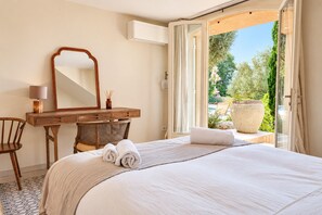 Une chambre d’hôtel comprenant un lit, un bureau, une chaise, un grand miroir et une porte ouverte donnant sur un balcon avec vue sur la verdure et un grand jardinière.