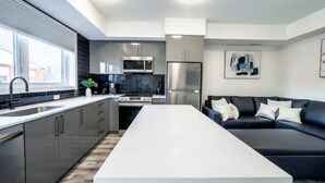 A modern kitchen with stainless steel appliances, a central island, and a black leather sofa in a living area. - Cozy 2BR Suite with Parking and 5 min Drive To Falls Sleeps 6! (Niagara Falls)