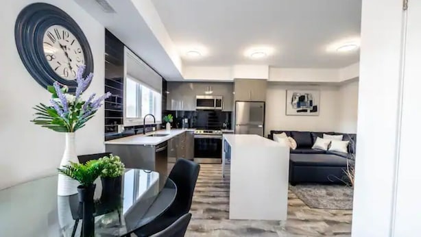 A modern kitchen with a central island, a dining area with a glass table, and a living area with a sofa. - Cozy 2BR Suite with Parking and 5 min Drive To Falls Sleeps 6! (Niagara Falls)