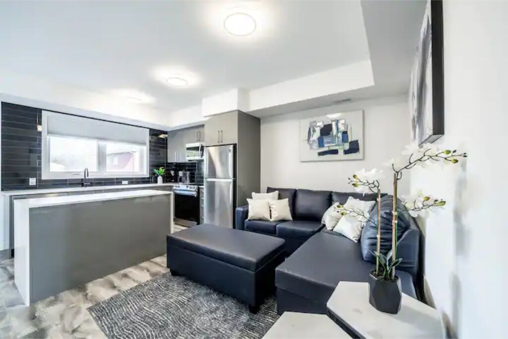 Una cocina moderna con una isla central, un área de comedor con un sofá y reposapiés, y un salón con un sofá y plantas decorativas.