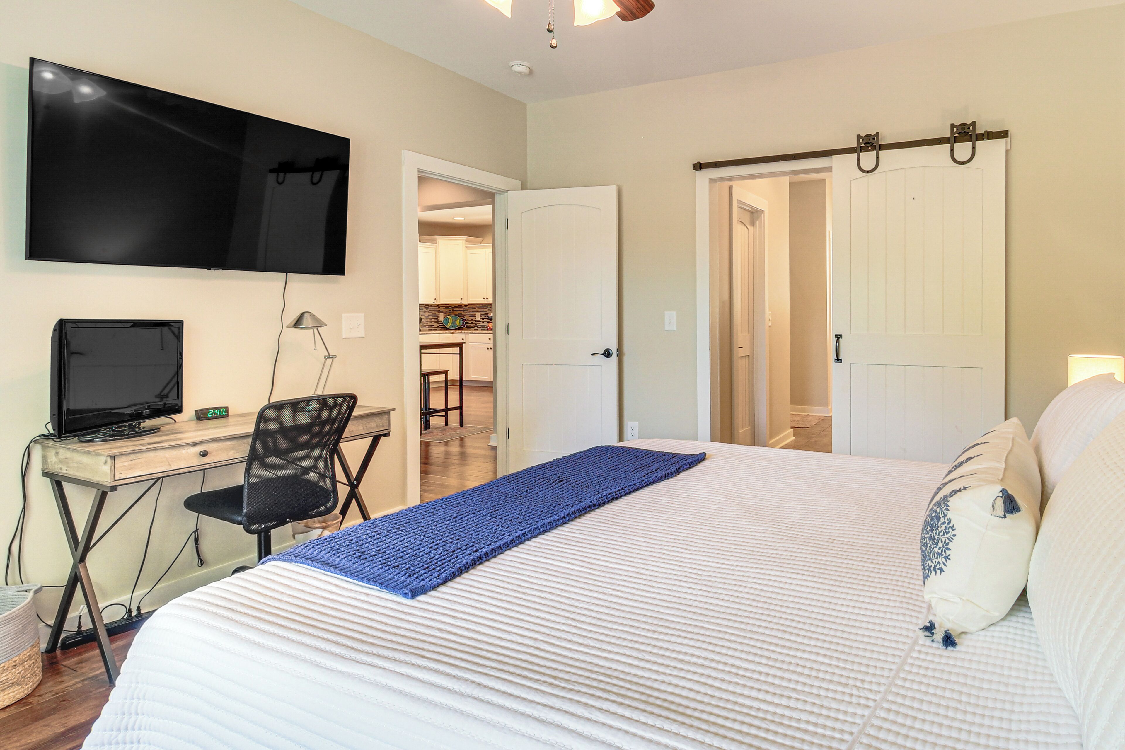 A bedroom with a bed, a desk with a computer, a TV mounted on the wall, and a door leading to another room.