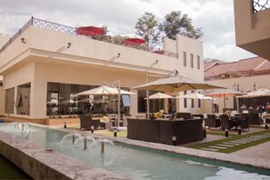 A pool area with lounge chairs and umbrellas, a water fountain, and a building with a balcony.