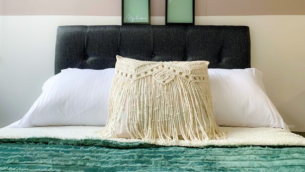 Una cama con un cojín de macramé, almohadas blancas y una manta verde.