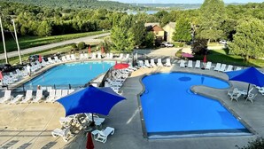 A pool area with two pools, surrounded by lounge chairs and umbrellas, set against a backdrop of trees and hills. - Hot Tub + Arcade -> Memorable Stay
 (Hollister)