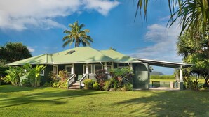 A green house with a porch, surrounded by lush greenery and a clear blue sky. - Beachfront Vacation Rental with AC throughout (Hanalei)