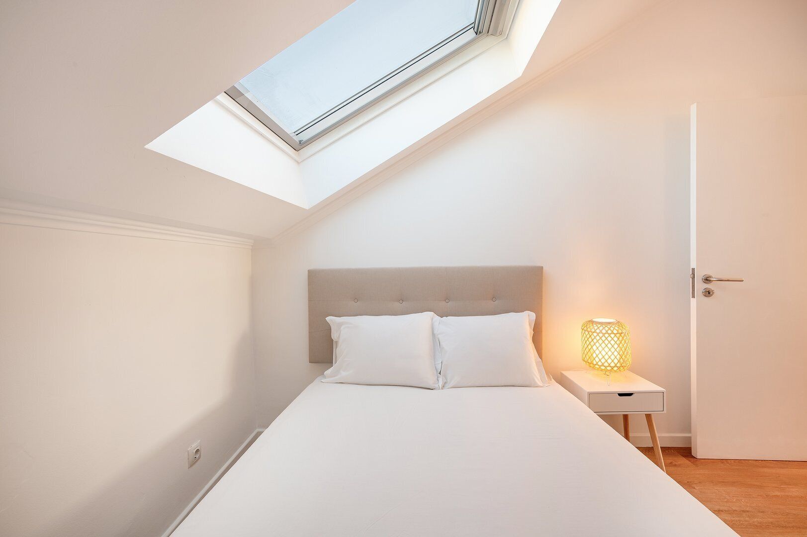 A bedroom with a skylight, a bed with white bedding, a nightstand with a lamp, and a door.