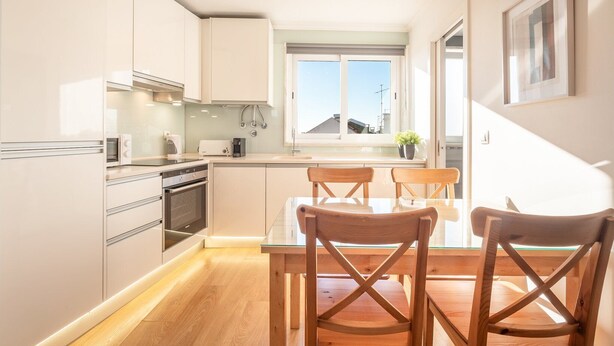 A modern kitchen with white cabinets, a dining table with wooden chairs, and a window with a view of buildings. - GuestReady - Sunny spacious in Belém (Lisbon)