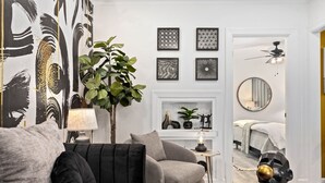 A living room with a grey sofa, a black armchair, a small side table with a lamp, and a decorative plant. There is a fireplace with decorative items and a bed in the background. - Designer Digs (Monrovia)