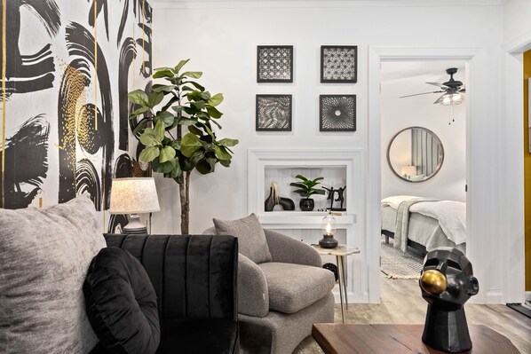 A living room with a grey sofa, a black armchair, a small side table with a lamp, and a decorative plant. There is a fireplace with decorative items and a bed in the background.