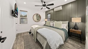A bedroom with a bed, a ceiling fan, a wall-mounted TV, a mirror, and an air conditioning unit. - Designer Digs (Monrovia)