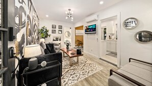 A modern living room with a sofa, coffee table, and TV. There is a kitchen area visible through an open door. - Designer Digs (Monrovia)