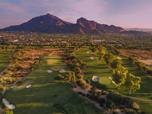 A golf course with bunkers and trees, surrounded by a residential area and mountains in the background.