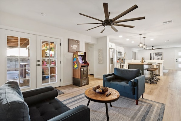 A modern living room with a sofa, armchair, and a coffee table. There is a ceiling fan, a kitchen in the background, and a game machine.