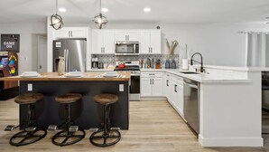 Uma cozinha moderna com armários brancos, uma ilha central com banquetas, eletrodomésticos de aço inoxidável e iluminação pendente.