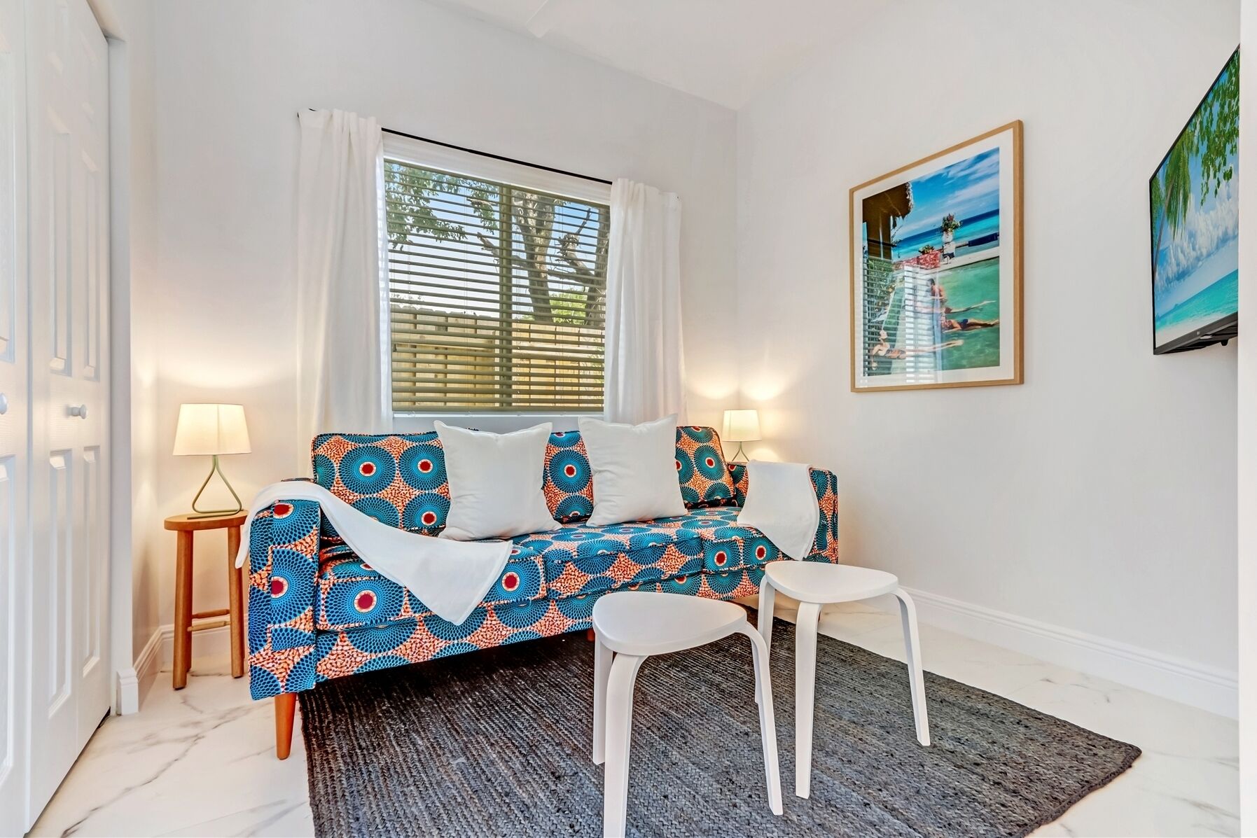 Een kamer met een gestreept bankje, twee witte stoelen, een kleine tafel en een raam met jaloezieën.