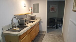 A compact kitchen with a sink, toaster, and wooden cabinets. A fire extinguisher is mounted on the wall, and a framed painting hangs above a table with chairs.