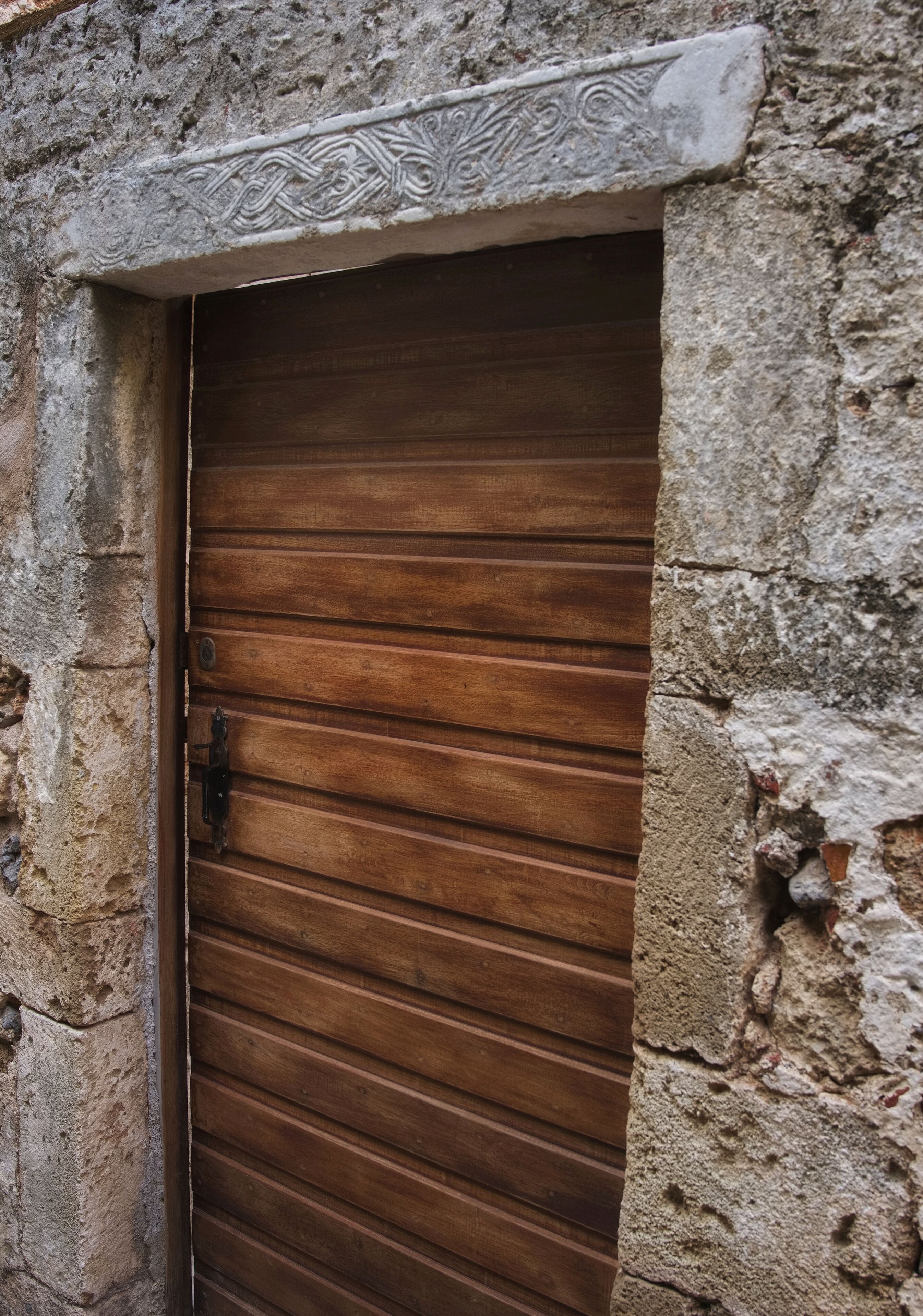 A wooden door with horizontal slats, set in a stone wall with a decorative stone lintel above it.