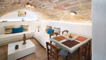 Una habitación acogedora con techo de piedra, un sofá blanco con cojines estampados, una mesa de comedor de madera con sillas, y una mesita blanca con un florero.