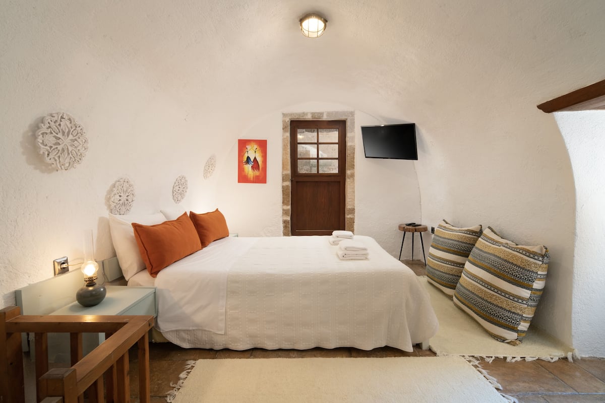 Ein Schlafzimmer mit einem Bett, Kissen, einem Nachttisch mit Lampe, einem an der Wand montierten Fernseher und einer Tür mit Fenster.