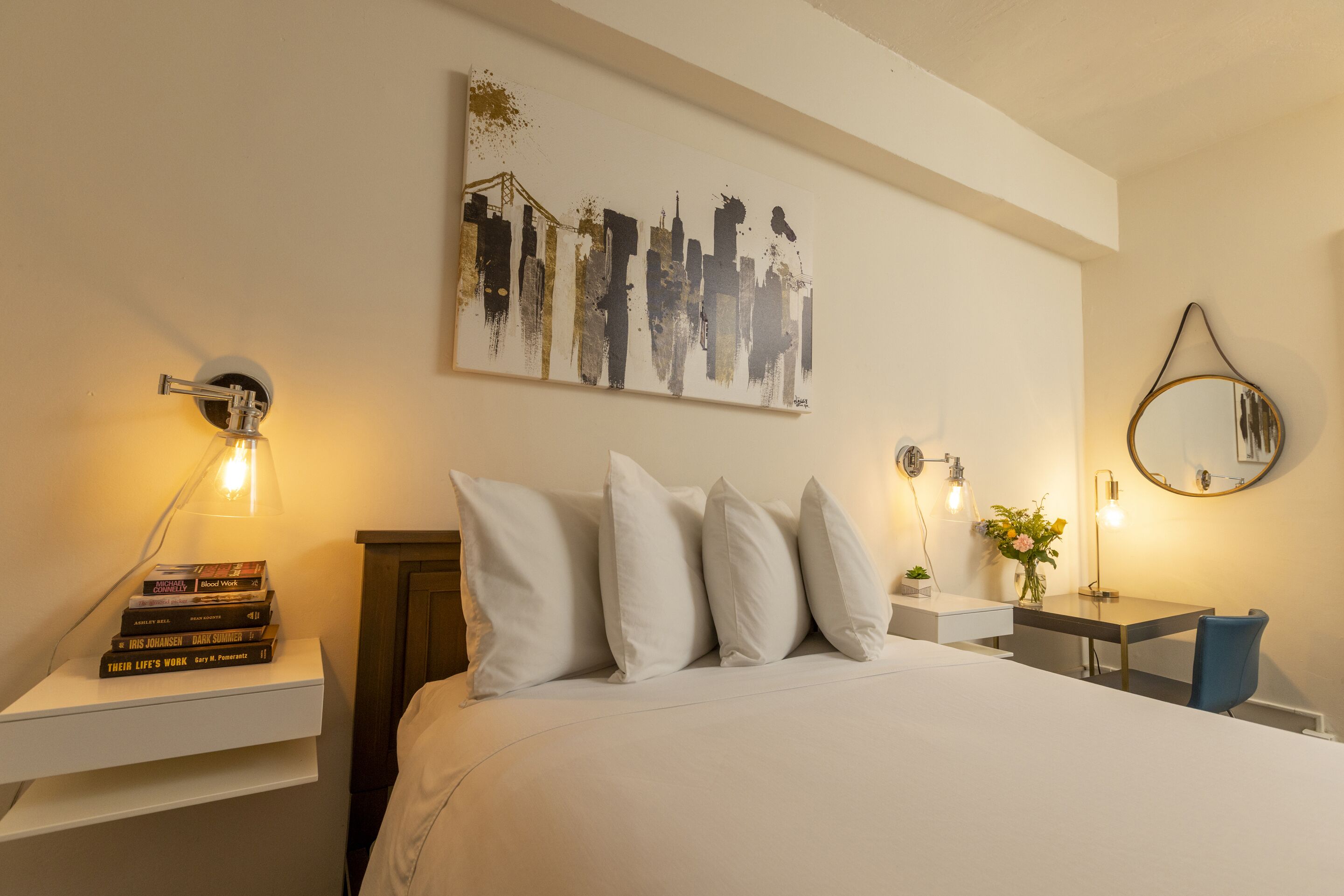 Un dormitorio ordenado con cama, almohadas, una mesita de noche con libros, una obra de arte en la pared, una lámpara de noche y un espejo.