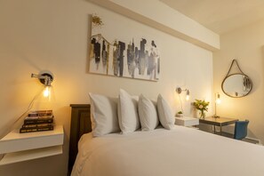 A neatly arranged bedroom with a bed, pillows, a bedside table with books, a wall-mounted artwork, a bedside lamp, and a mirror.