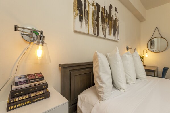 A neatly made bed with white bedding and pillows, a bedside table with a stack of books, a wall-mounted abstract painting, and a wall-mounted mirror.