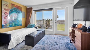 Ein Schlafzimmer mit einem Bett, einer Bank, einem Fernseher, einer Holzkommode, einem großen Gemälde und einer Glastür zu einem Balkon.