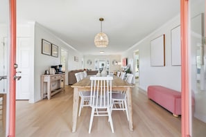 Un vaste espace dédié à la salle à manger, avec une table en bois et des chaises blanches, un luminaire suspendu et un banc à l’arrière-plan.