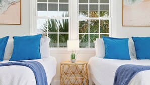 A room with two beds, each with blue and white bedding, a small round table with a plant, and a view of palm trees through large windows.