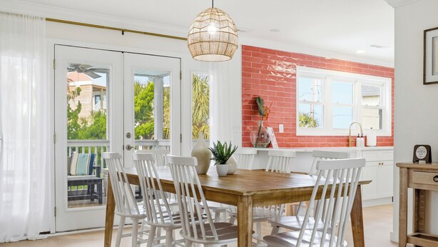 A dining area with a wooden table and white chairs, a hanging light fixture, and a red brick wall.