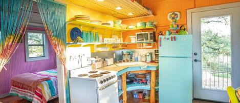Cocina compacta con estufa, microondas y refrigerador, una cama con manta a rayas coloridas, y ventana con vista a árboles.