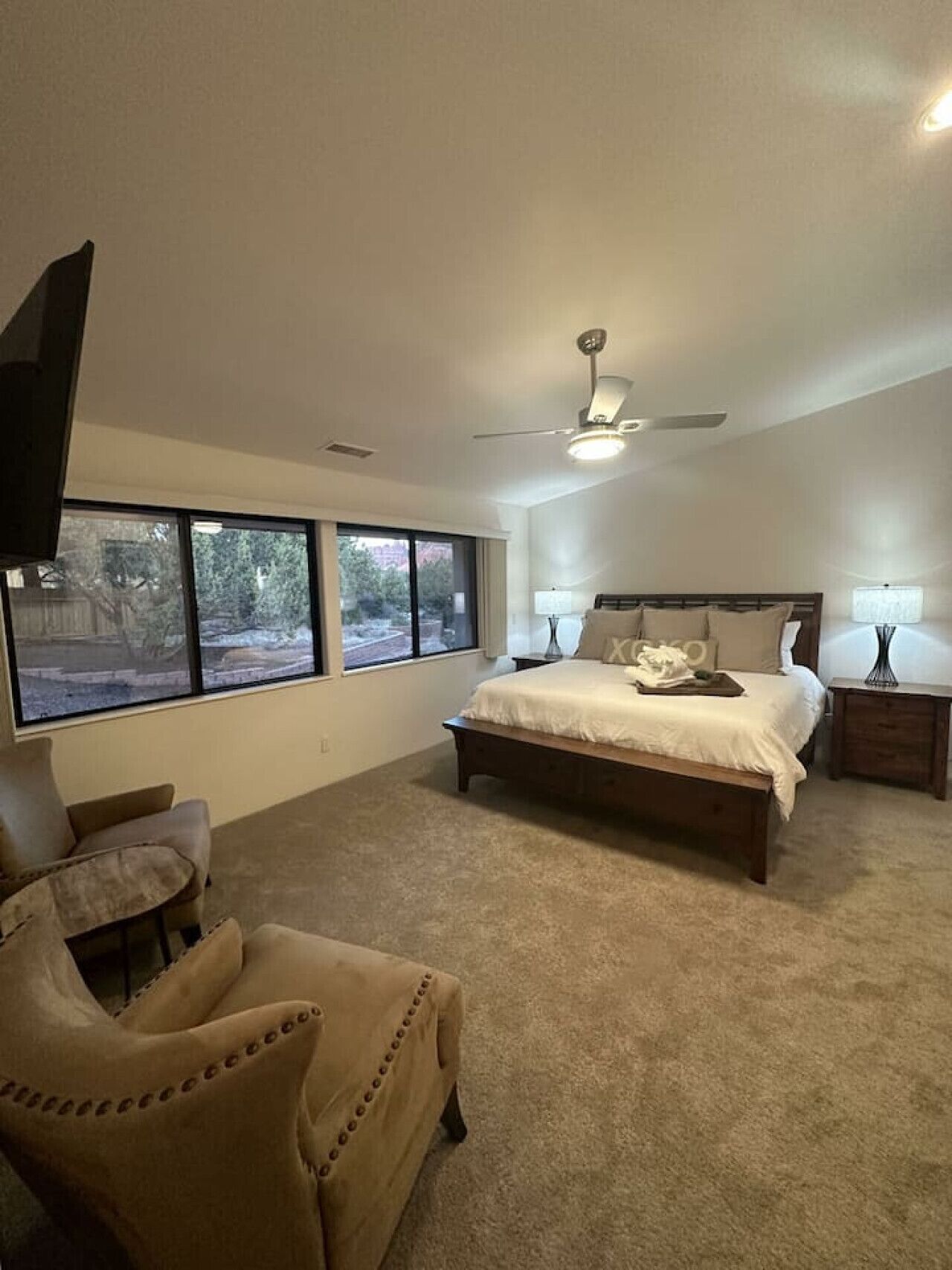 A bedroom with a large bed, two bedside tables, a ceiling fan, and a TV mounted on the wall.