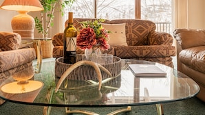 Uma sala de estar com uma mesa de centro de vidro, sofás estampados, um abajur, um vaso com flores e uma garrafa.
