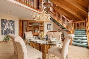 A dining area with a glass table, cushioned chairs, a wooden staircase, and a chandelier.