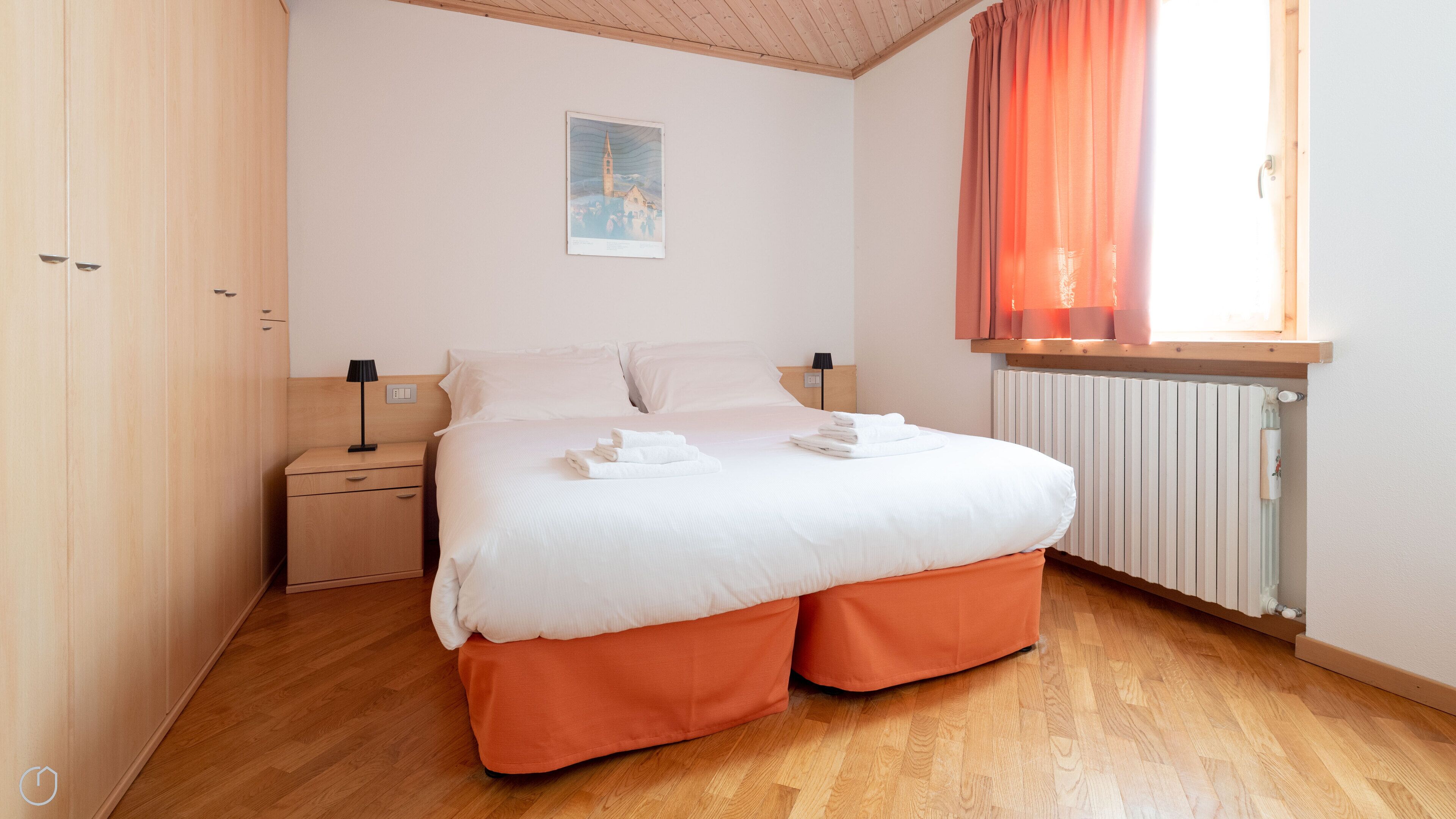Cama bem arrumada com lençóis brancos e pés de apoio laranja, mesa de cabeceira com abajur, e janela com cortinas laranja.