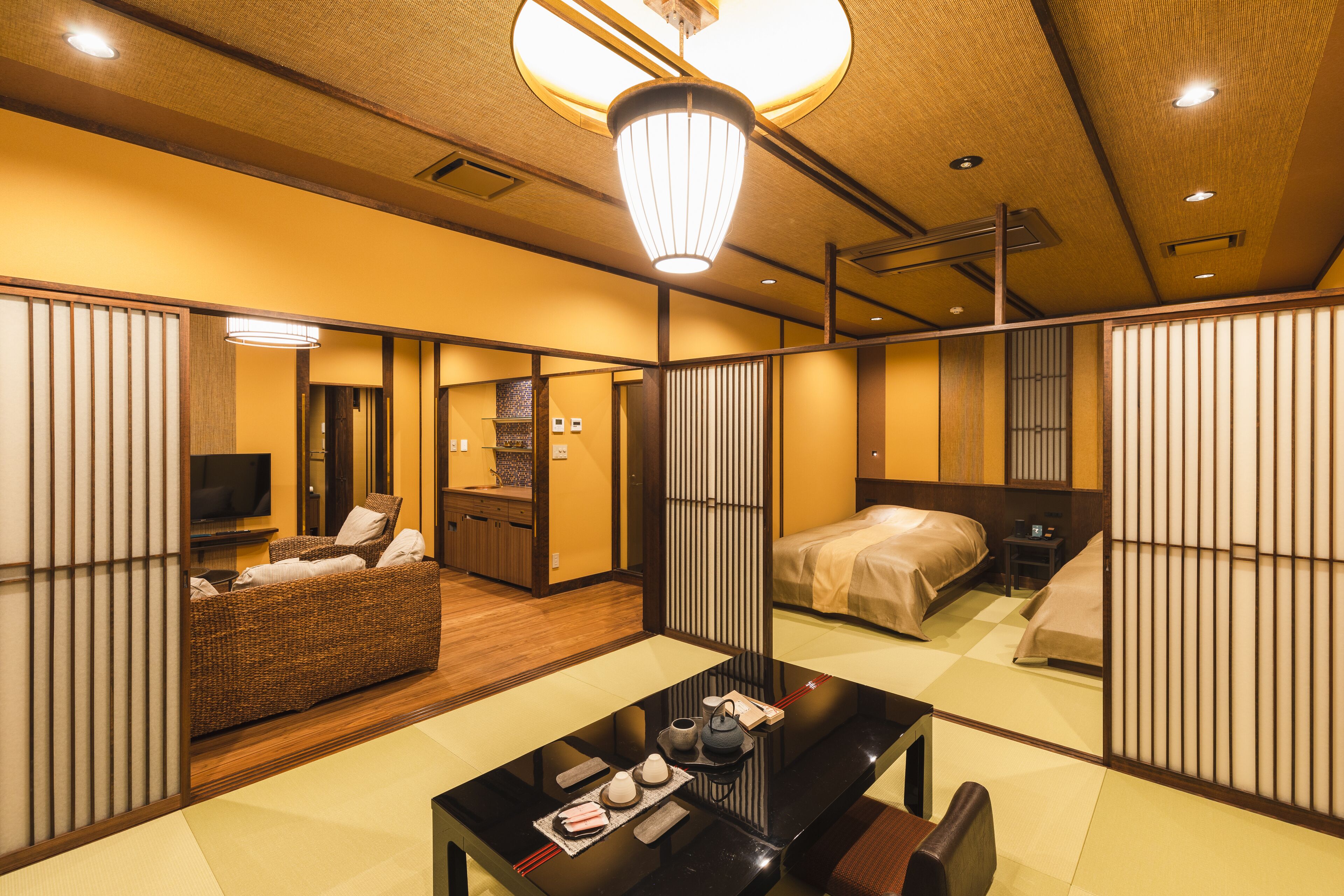 A traditional Japanese-style hotel room with tatami flooring, a low table with tea set, a sofa, a bed, and sliding doors.