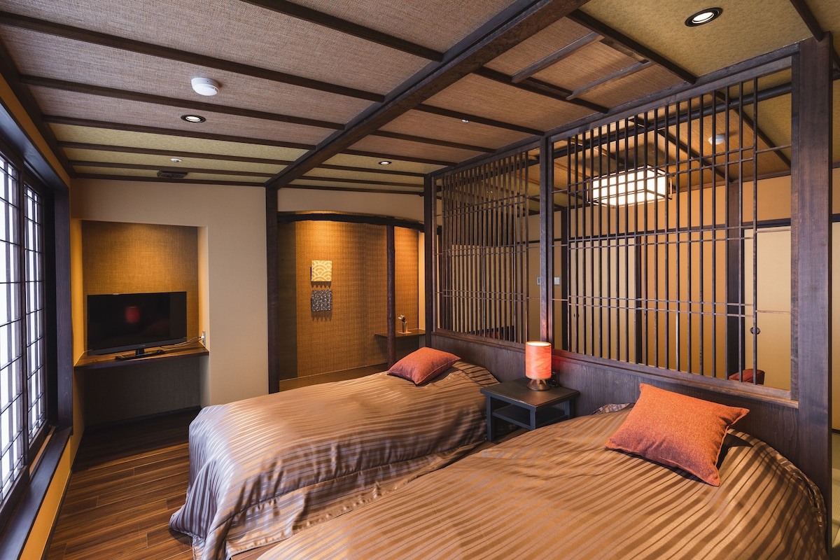 A traditional Japanese-style room with tatami flooring, two beds with striped bedding, a television on a low table, and a sliding door with a decorative screen.