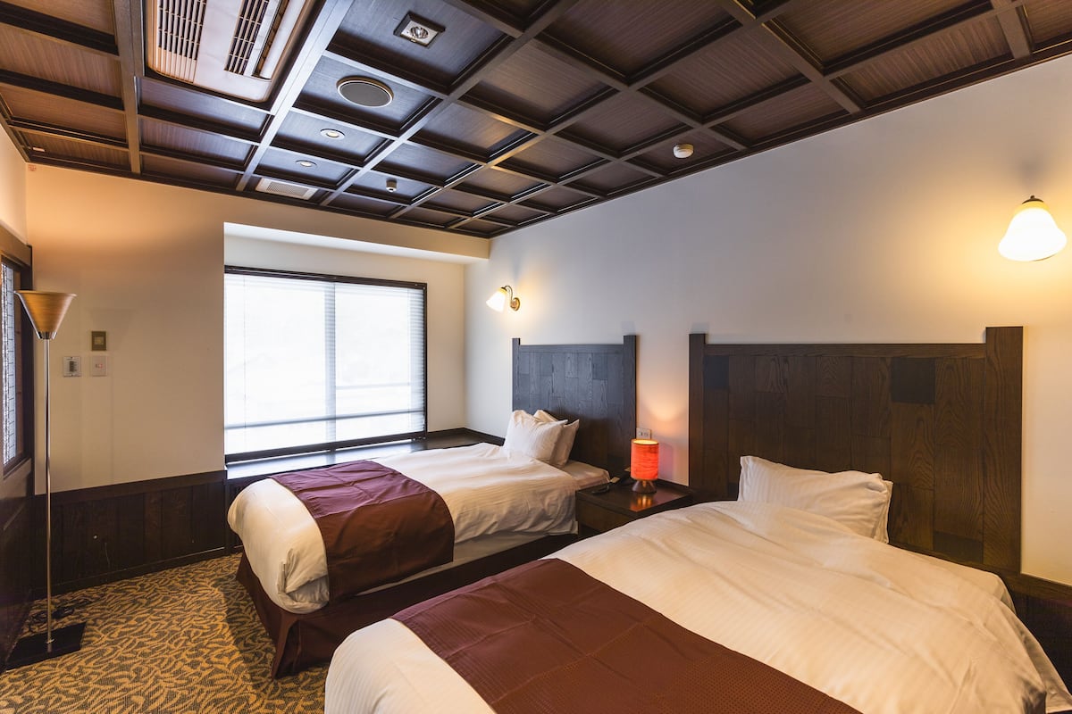 A hotel room with two beds, a window, a ceiling with wooden beams, and a wall-mounted lamp.