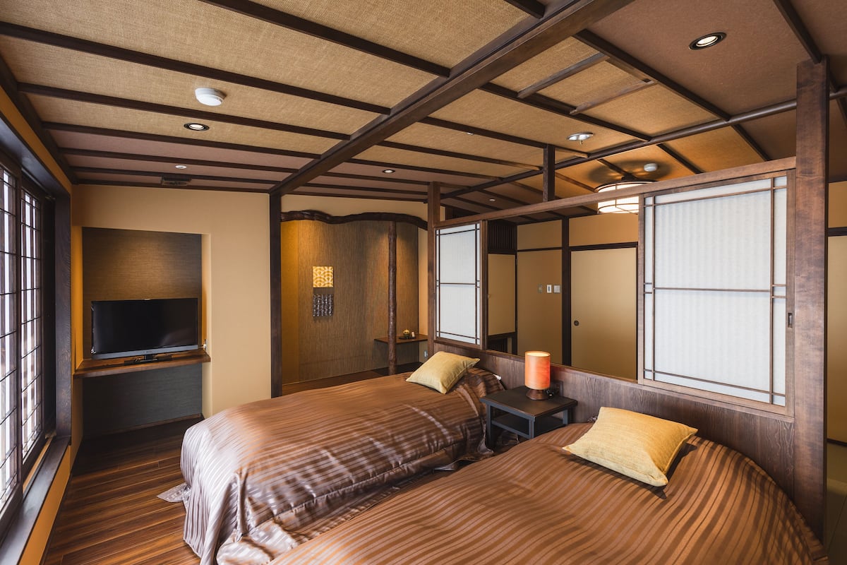 A traditional Japanese-style room with tatami flooring, a television on a low shelf, a bed with a wooden headboard, and a sliding shoji screen door.