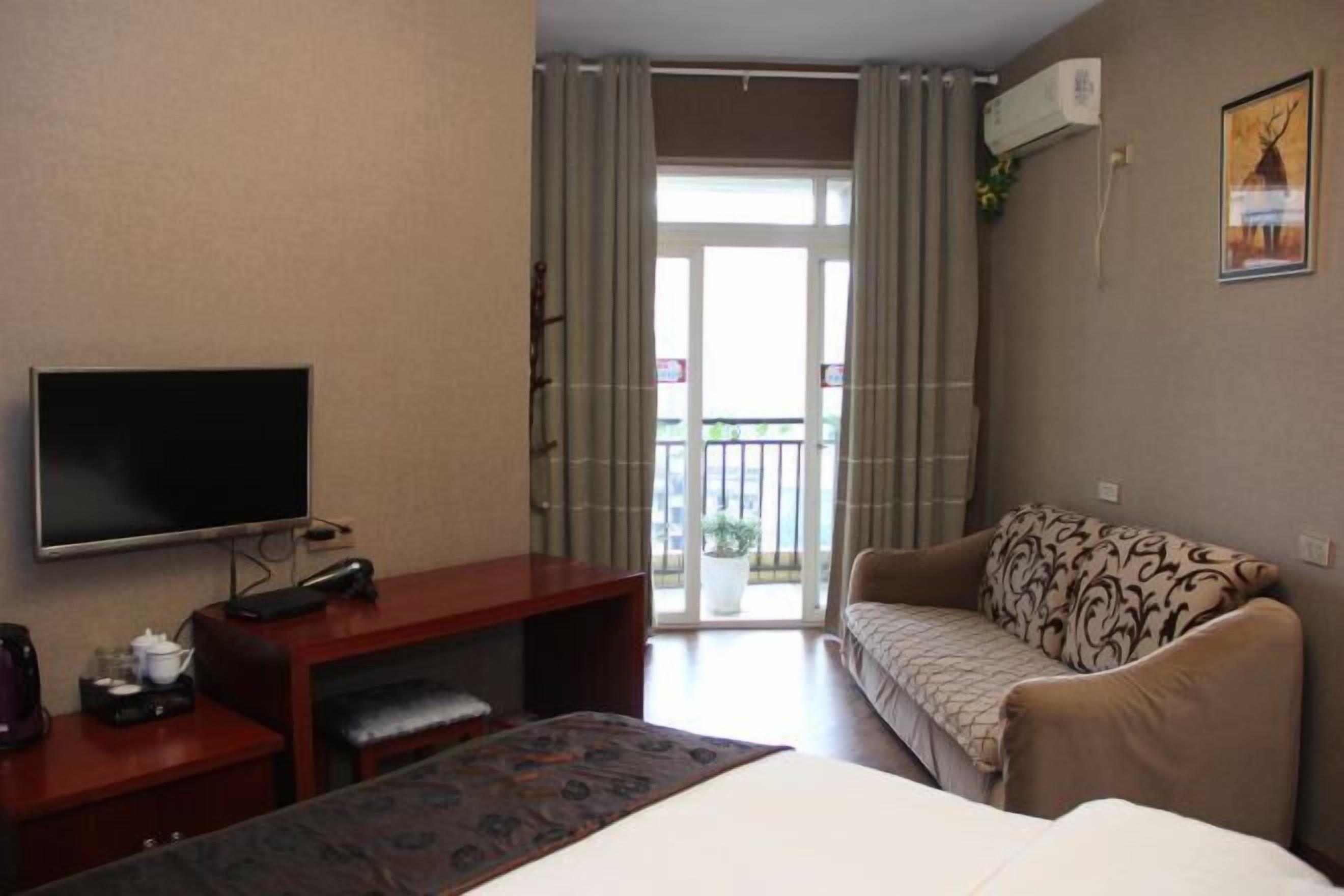 A hotel room with a bed, a desk with a computer, a television, a sofa, and a balcony with curtains.