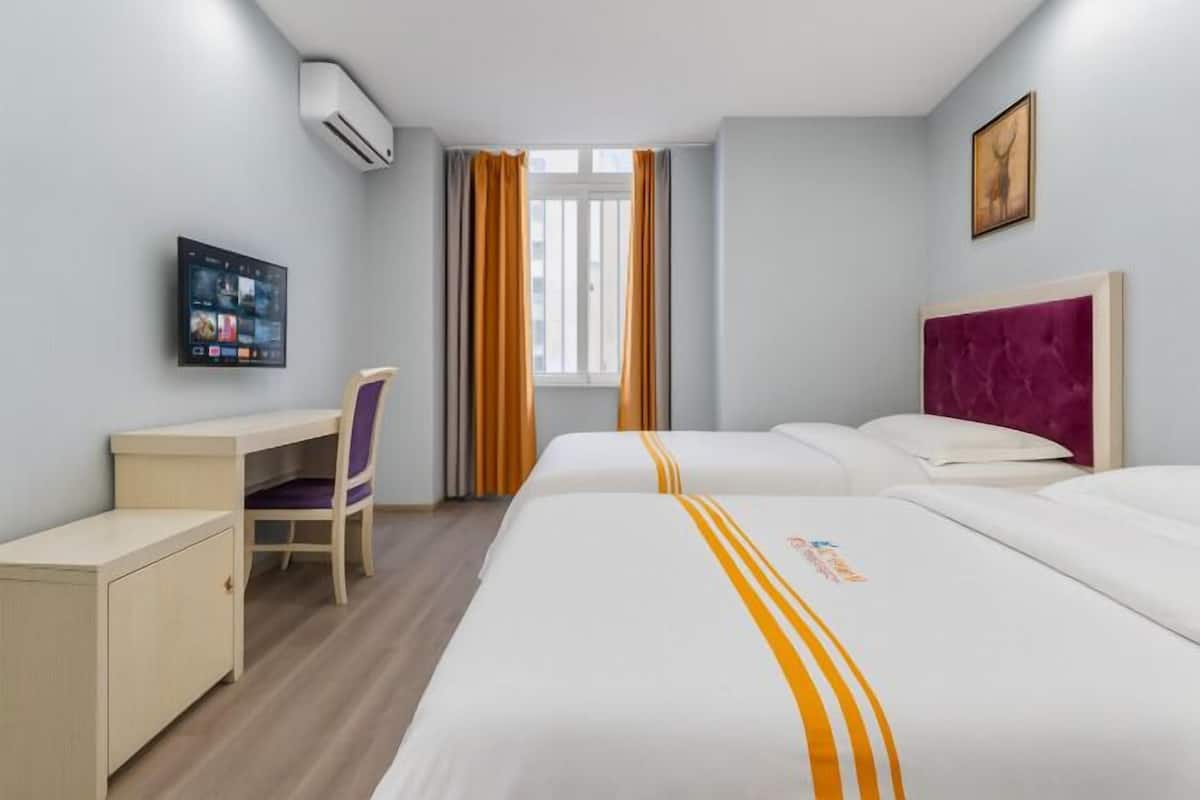 A hotel room with two beds, a desk, a chair, an air conditioning unit, and a window with curtains.