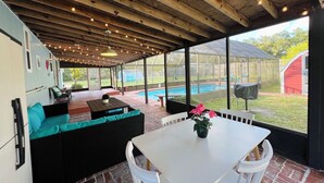 A covered patio area with a white table and chairs, a black sofa with teal cushions, and a view of a swimming pool and a red shed. - Serene Home with PRIVATE Pool - Near Beaches - Dog Friendly - Firepit&Large Yard (Freeport)