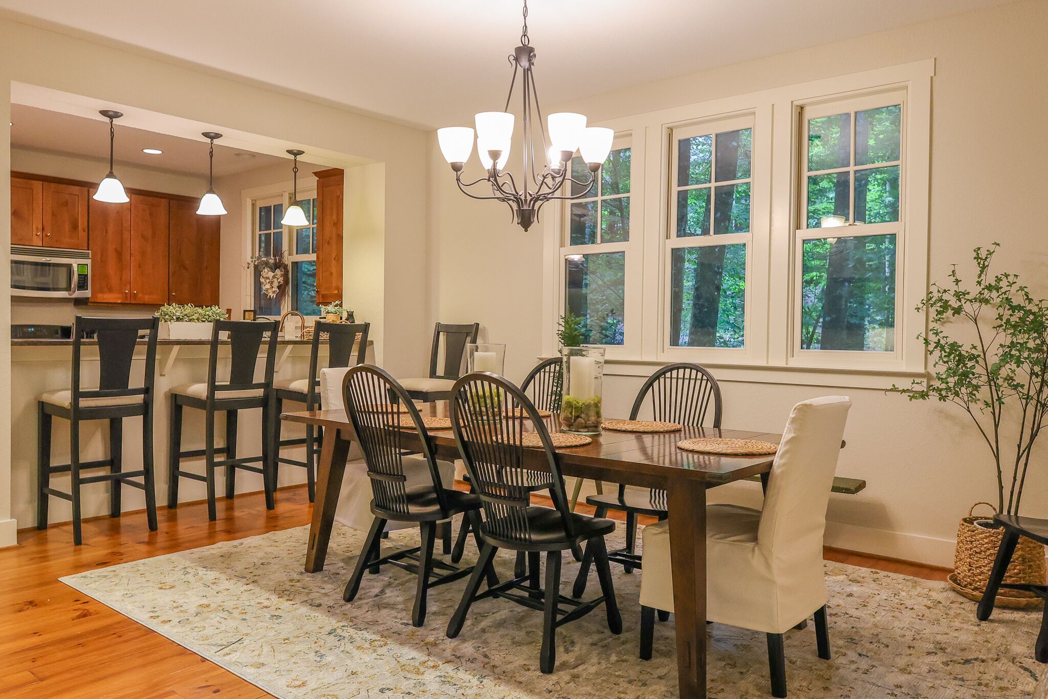 A dining area with a wooden table and chairs, a chandelier, and a view of the outdoors through large windows.