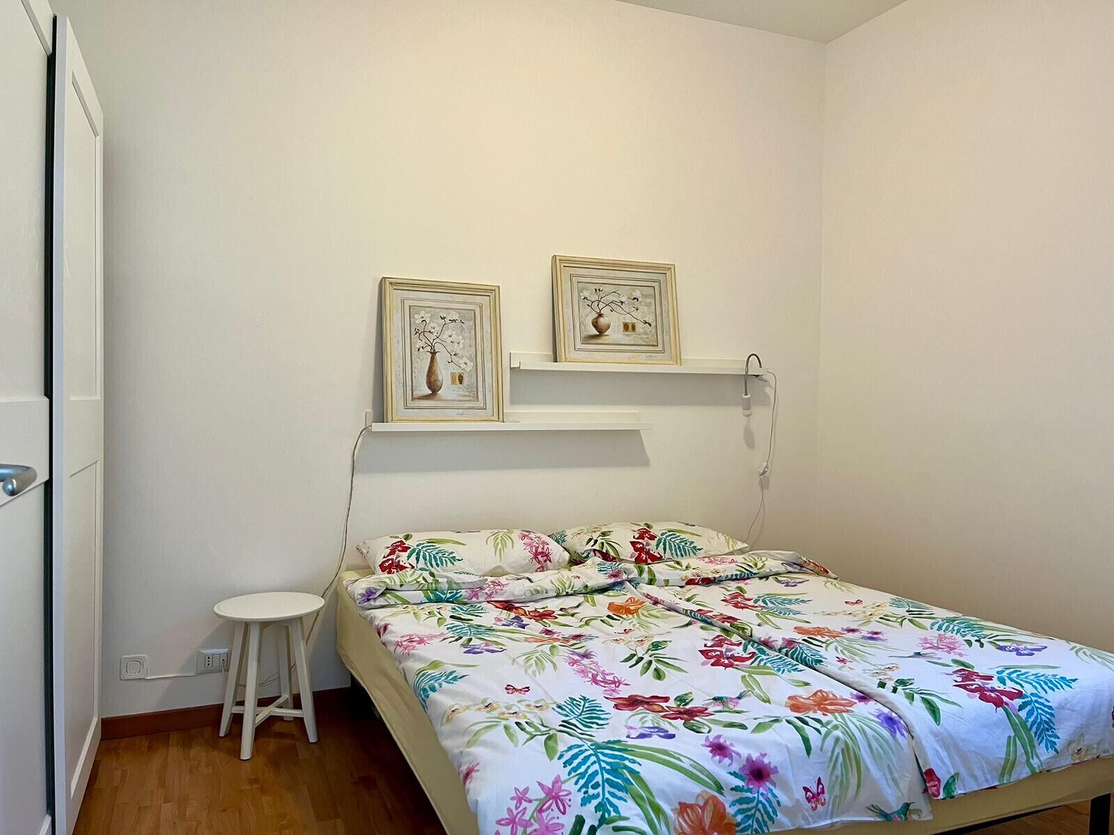 Un dormitorio pequeño y ordenado con una cama individual, un taburete blanco y dos cuadros en la pared.
