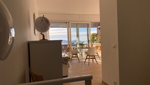 A room with a wooden floor, a white desk, a globe on a shelf, and a balcony with a view of the sea.