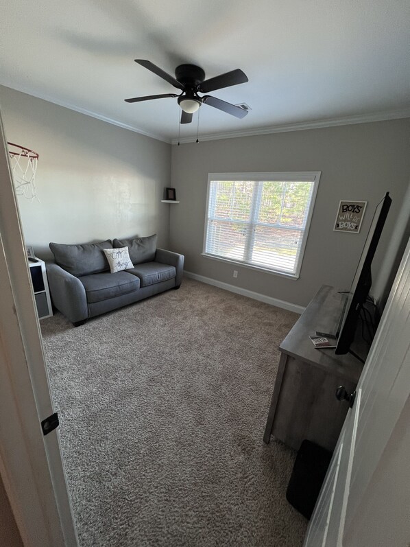 A living room with a grey sofa, a ceiling fan, a window with blinds, and a small shelf with a picture frame.