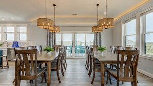 A dining area with a long wooden table and chairs, two hanging light fixtures, and a view through large windows. - Happy As a Clam  | 230 ft to the beach | Private Pool, Hot Tub, Dog Friendly (Virginia Beach)